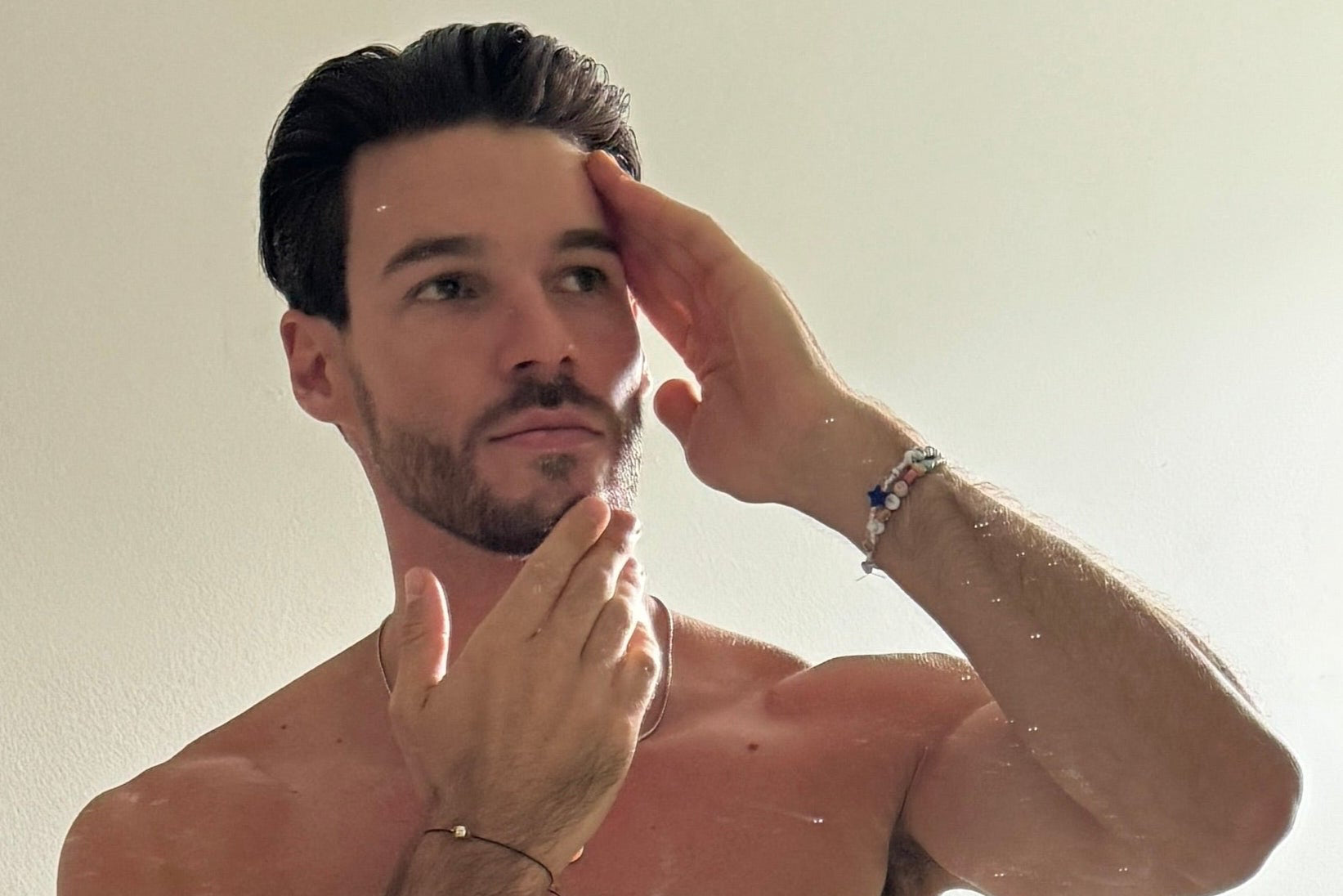 Man in a bathroom looking at himself in the mirror, touching his face.